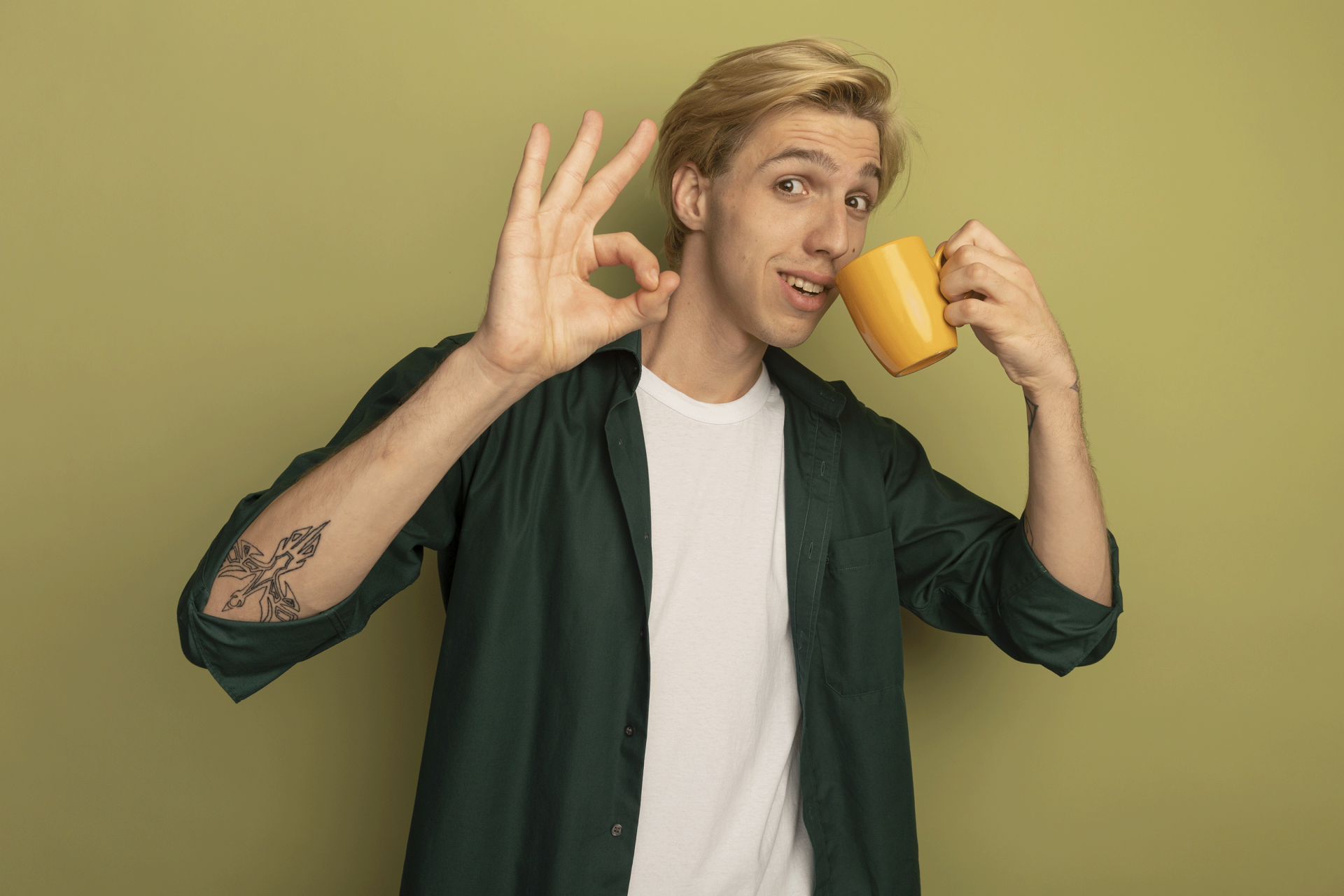 pleased-young-blonde-guy-wearing-green-t-shirt-drinks-tea-showing-okay-gesture